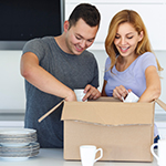 This image shows a young and in love smiling couple unpacking boxes in their new home. They are in the kitchen. We can see plates on the table and they taking cups from the box. In the background we can see kitchen elements.